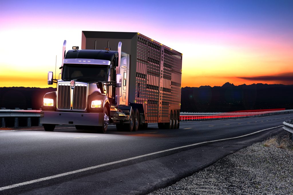 A black Kenworth truck driving down the road just after sunset with a purple, yellow, and orange sky behind it.