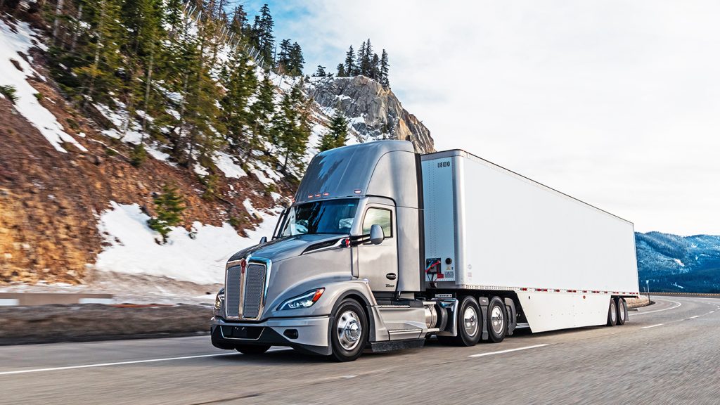 A silver Kenworth truck driving on the road with snow spotted mountains and trees behind it.