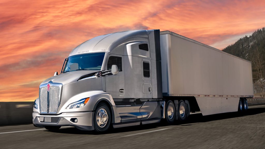 A silver Kenworth sleeper cab driving down the road with pink and orange clouds behind it.