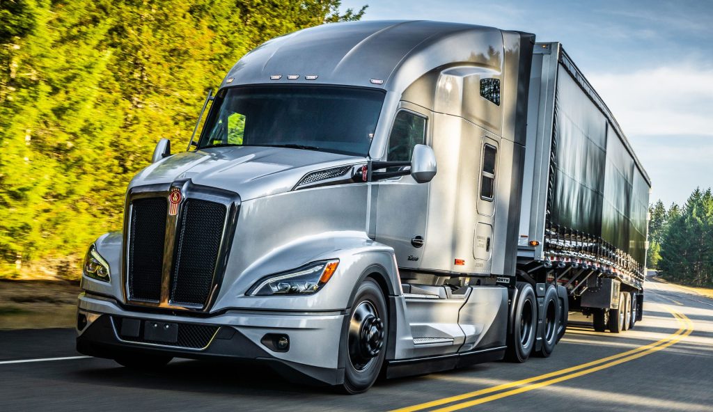 A silver Kenworth semi truck driving on a road surrounded by trees.